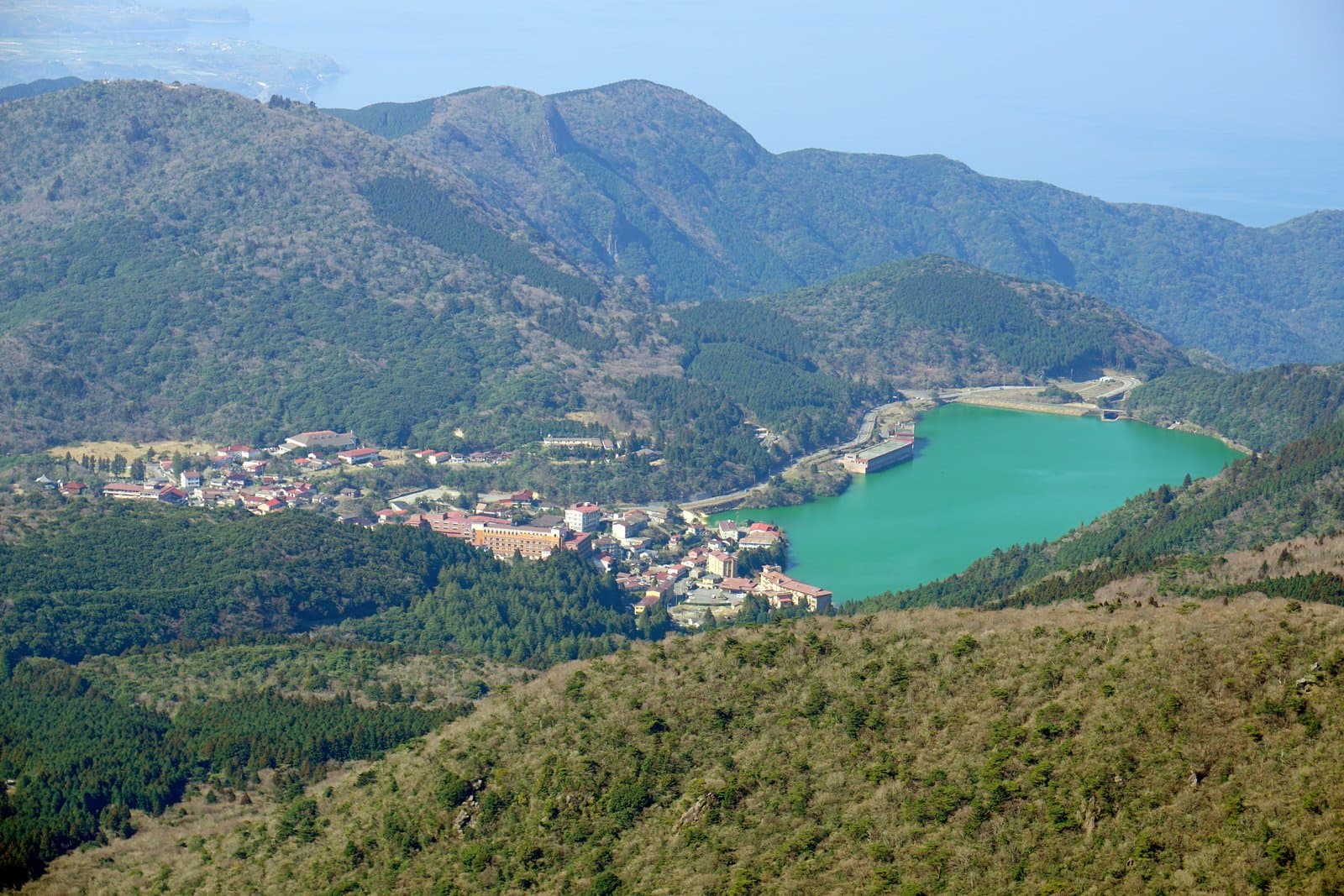 A hot spring town beside a turquoise reservoir among wooded mountains, seen from a peak