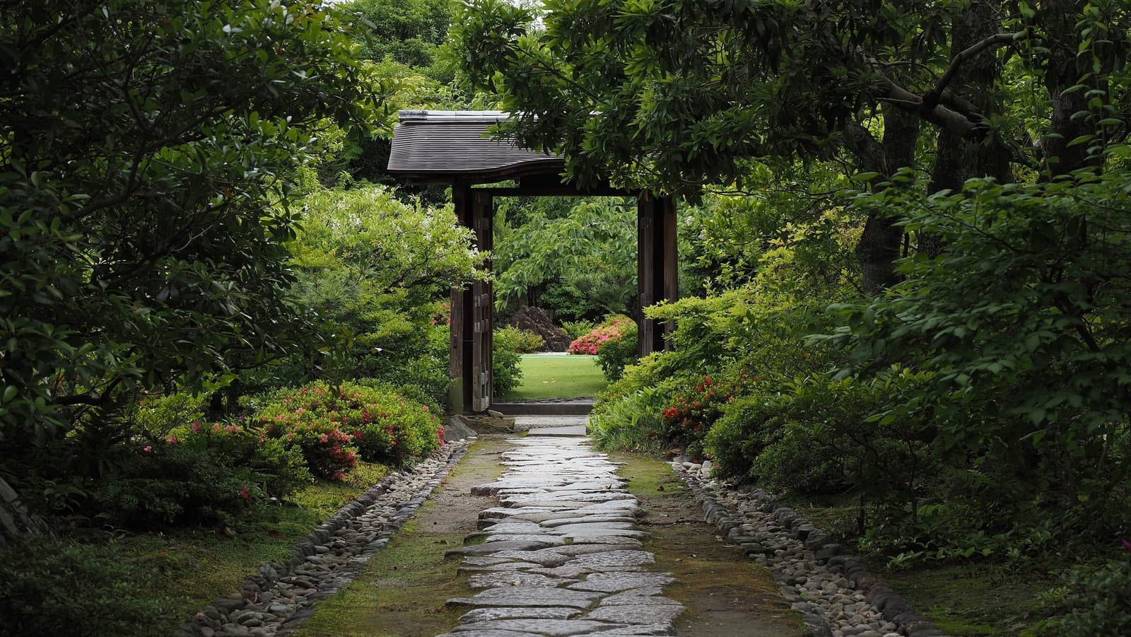A stone-paved garden path leading through fresh green trees and azaleas to a small roofed gate