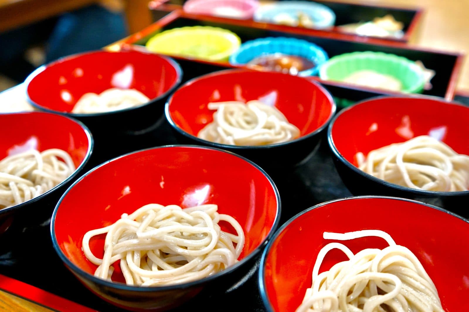 Small red lacquer bowls each holding a mouthful of soba, with side dishes behind