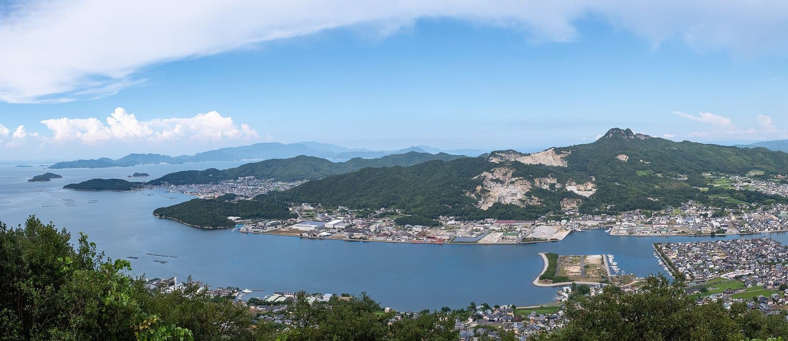 A bay with islands and a town below forested hills, seen from a mountain top