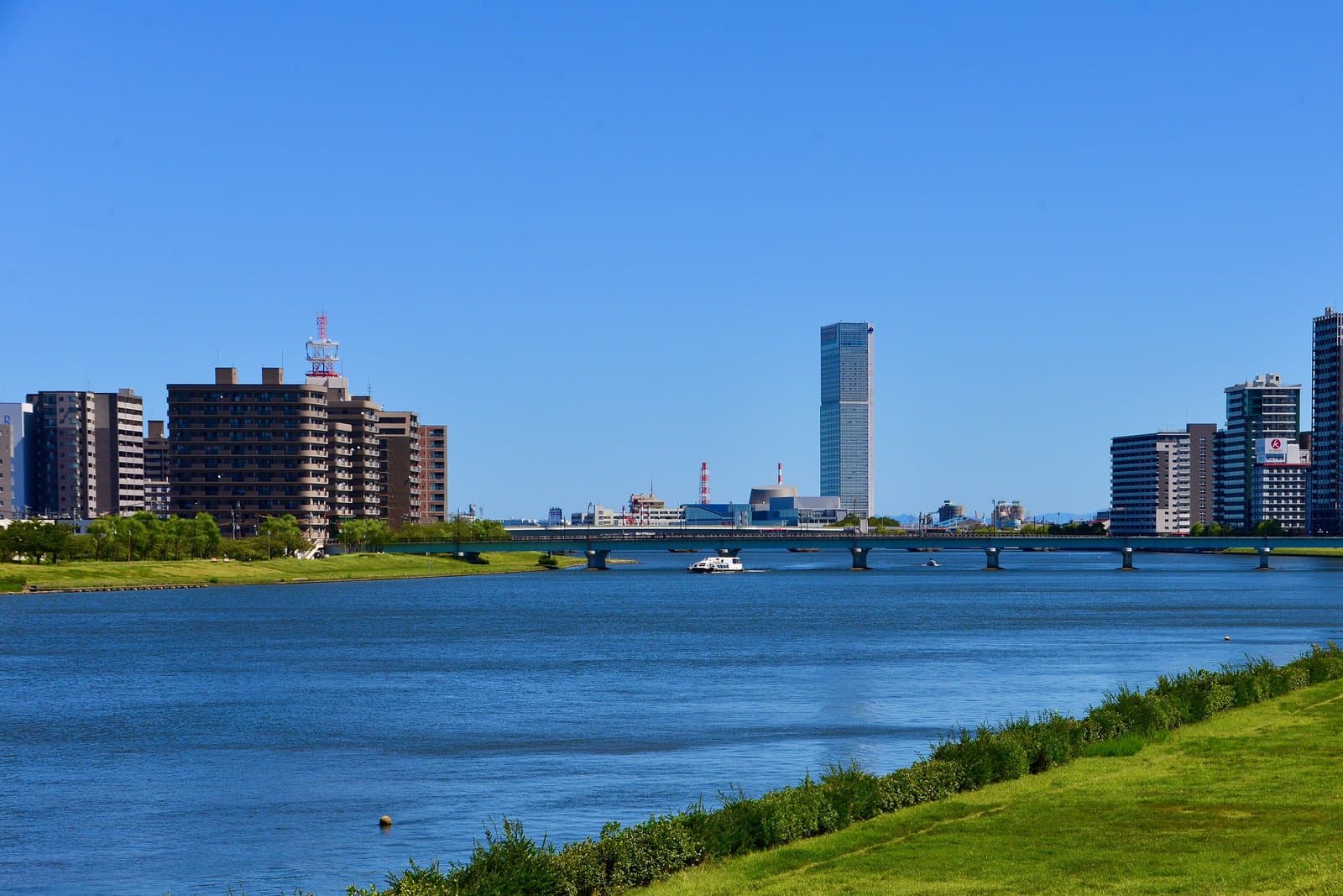 A wide river curving past a grassy embankment, with city buildings and a bridge in the distance under a blue sky