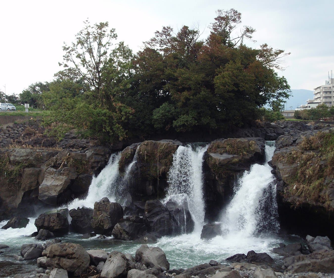 Several white waterfalls pouring over a dark lava cliff into a rocky river, with trees above