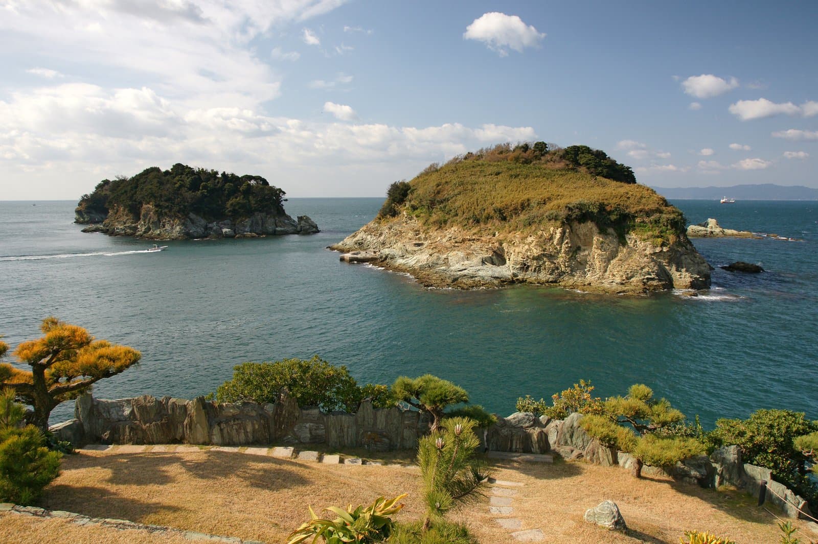 Two rocky islands in a blue-green sea, seen over a stone wall and pines at the edge of a garden