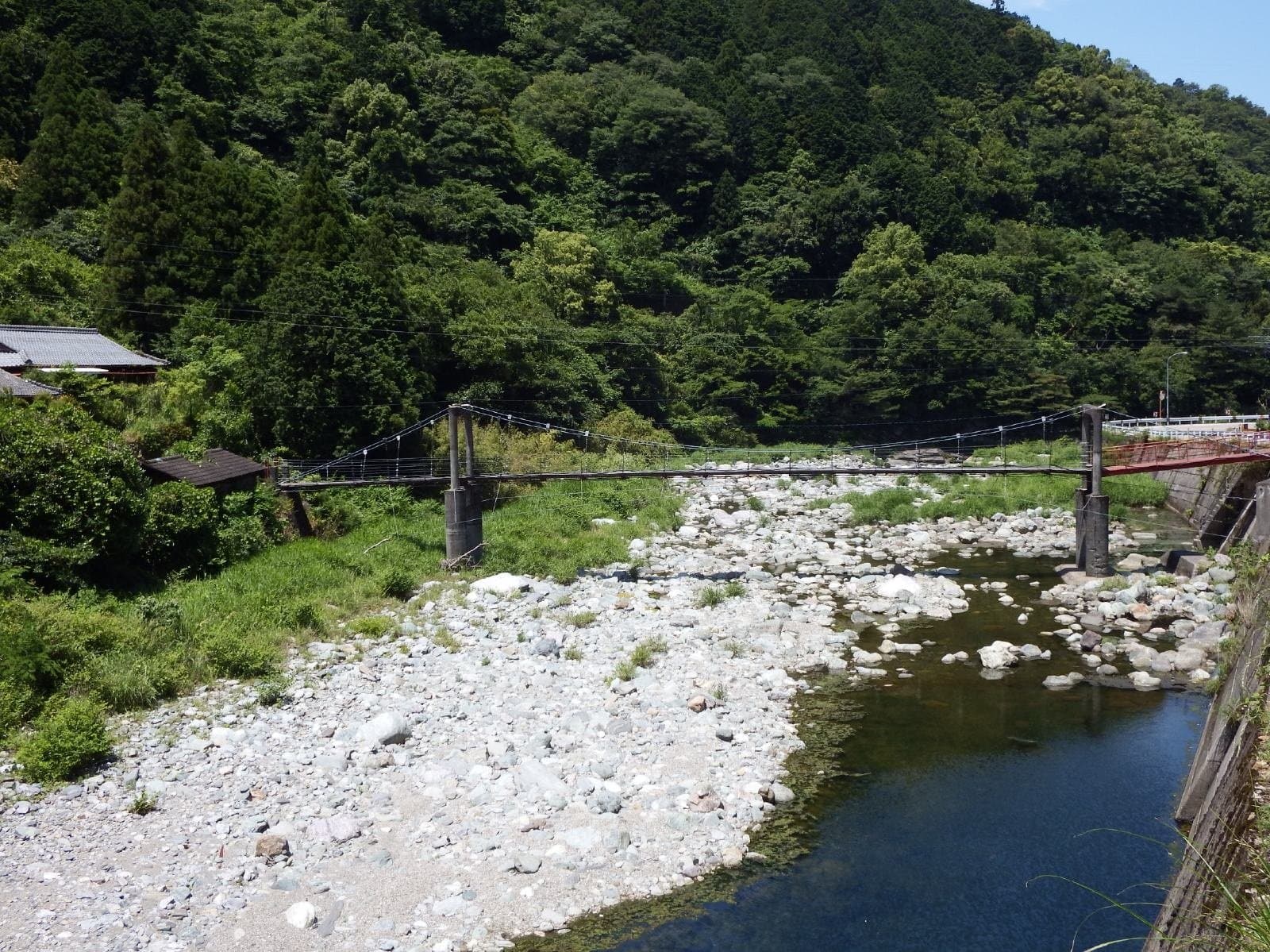 A small suspension bridge over a clear river with a wide bed of white stones, surrounded by green forested mountains