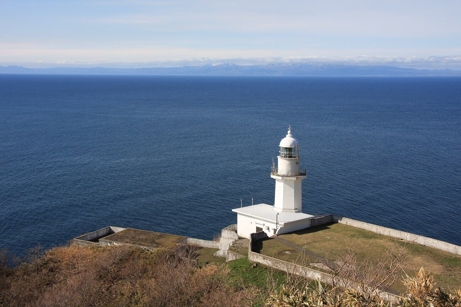 A white lighthouse on a clifftop above a deep blue sea, with distant mountains on the horizon