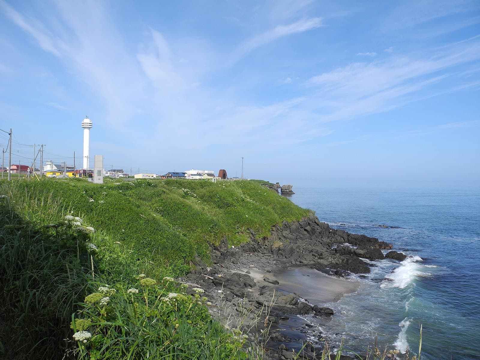A grassy clifftop above a rocky shore and calm sea, with a white tower and low buildings on the cape