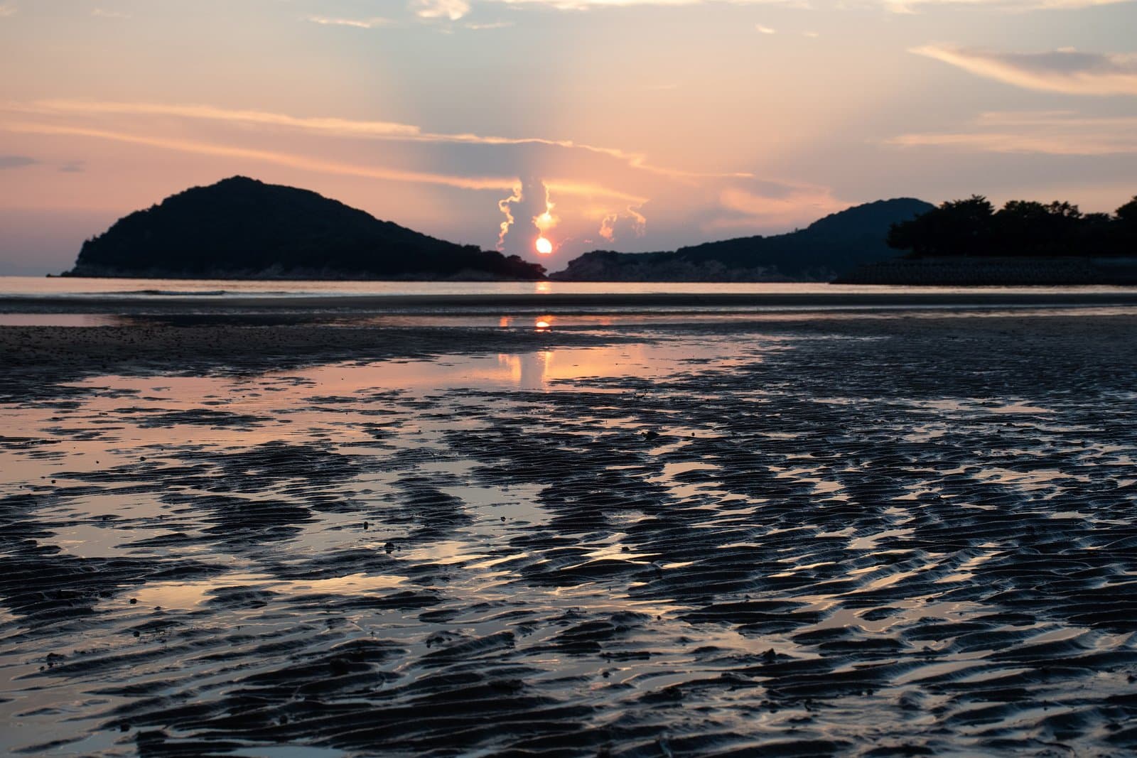 濡れた干潟の向こう、2つの丘の間に沈む夕日