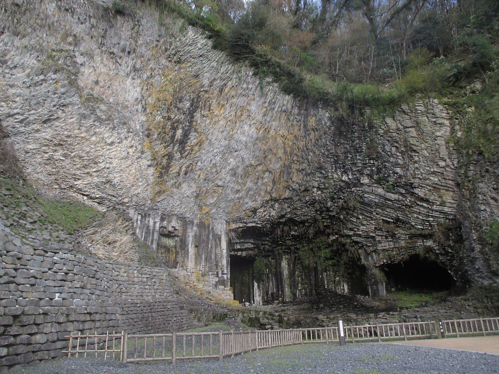 A tall cliff of columnar basalt above a cave opening, with a low wooden fence in front