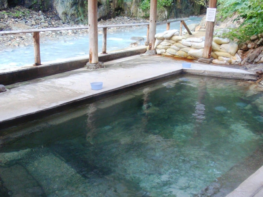 A clear open-air bath under a wooden roof beside a rocky mountain river
