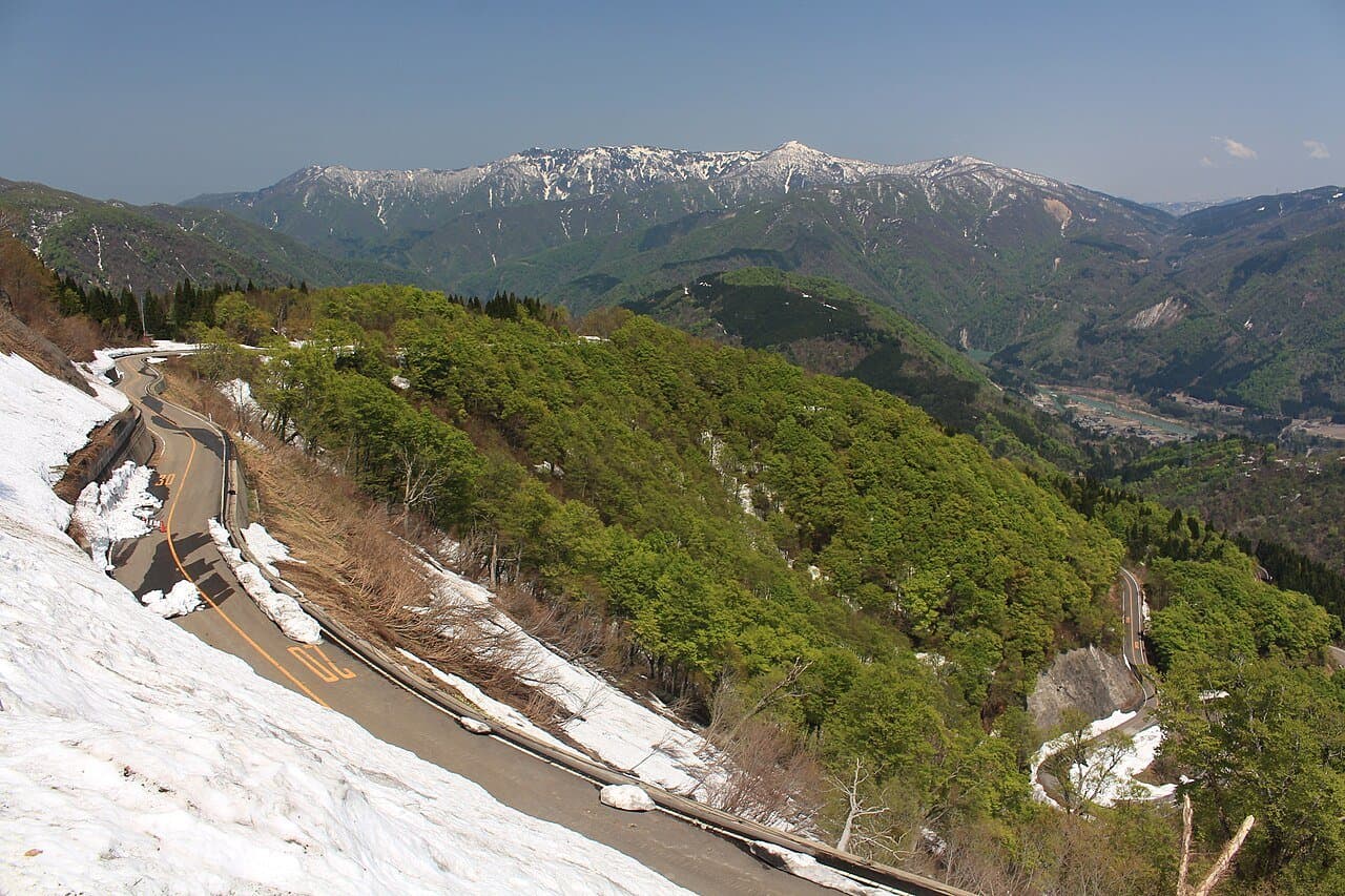 A mountain road winding along a snowy slope above fresh green forest, with snow-streaked peaks beyond