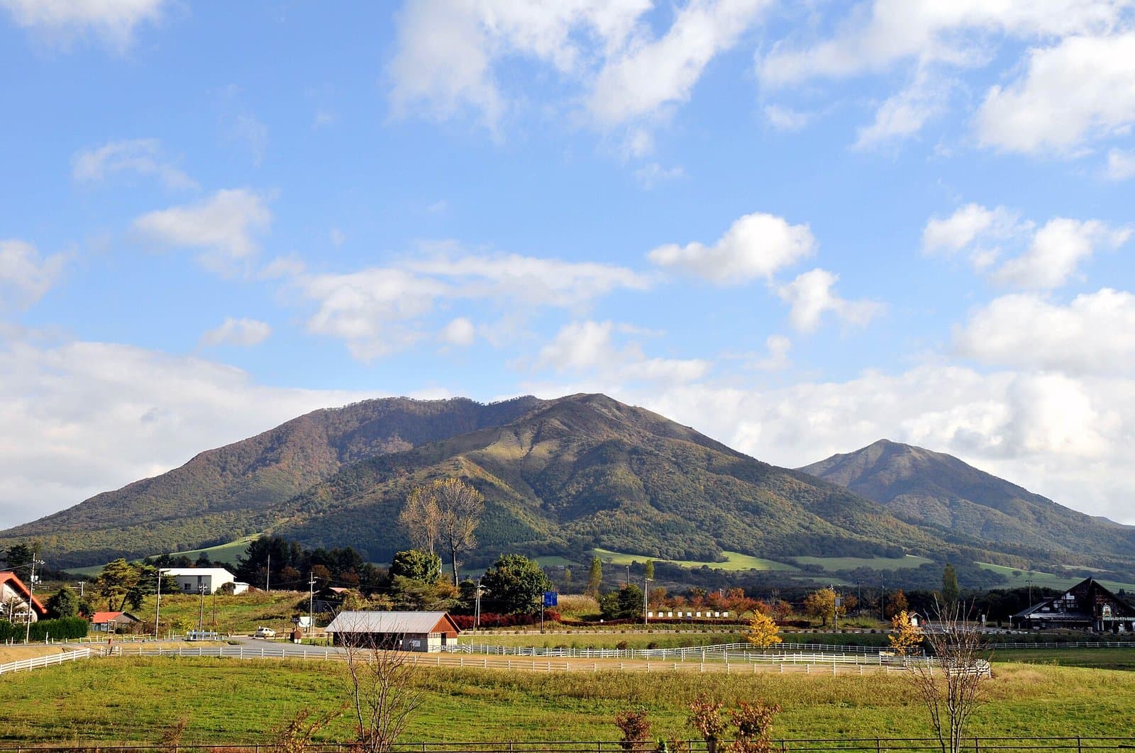 Green mountains rising beyond meadows and farm buildings under a blue sky with clouds