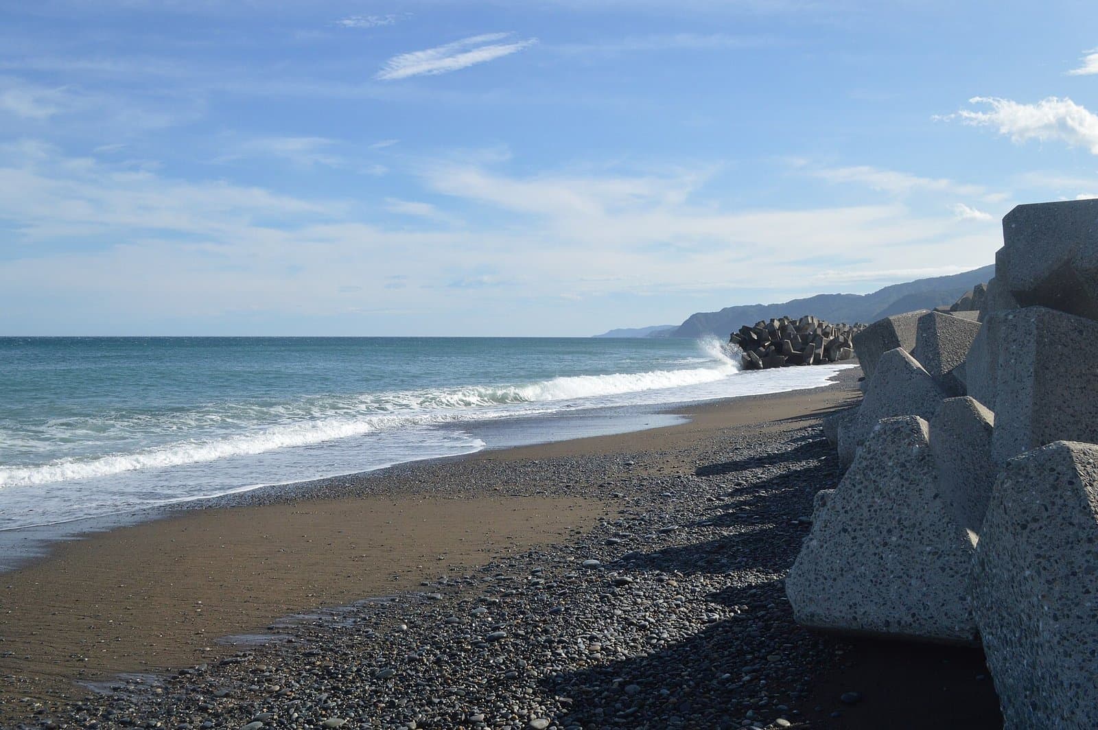 Waves breaking on a beach of small dark pebbles, with concrete wave-breaking blocks on the right