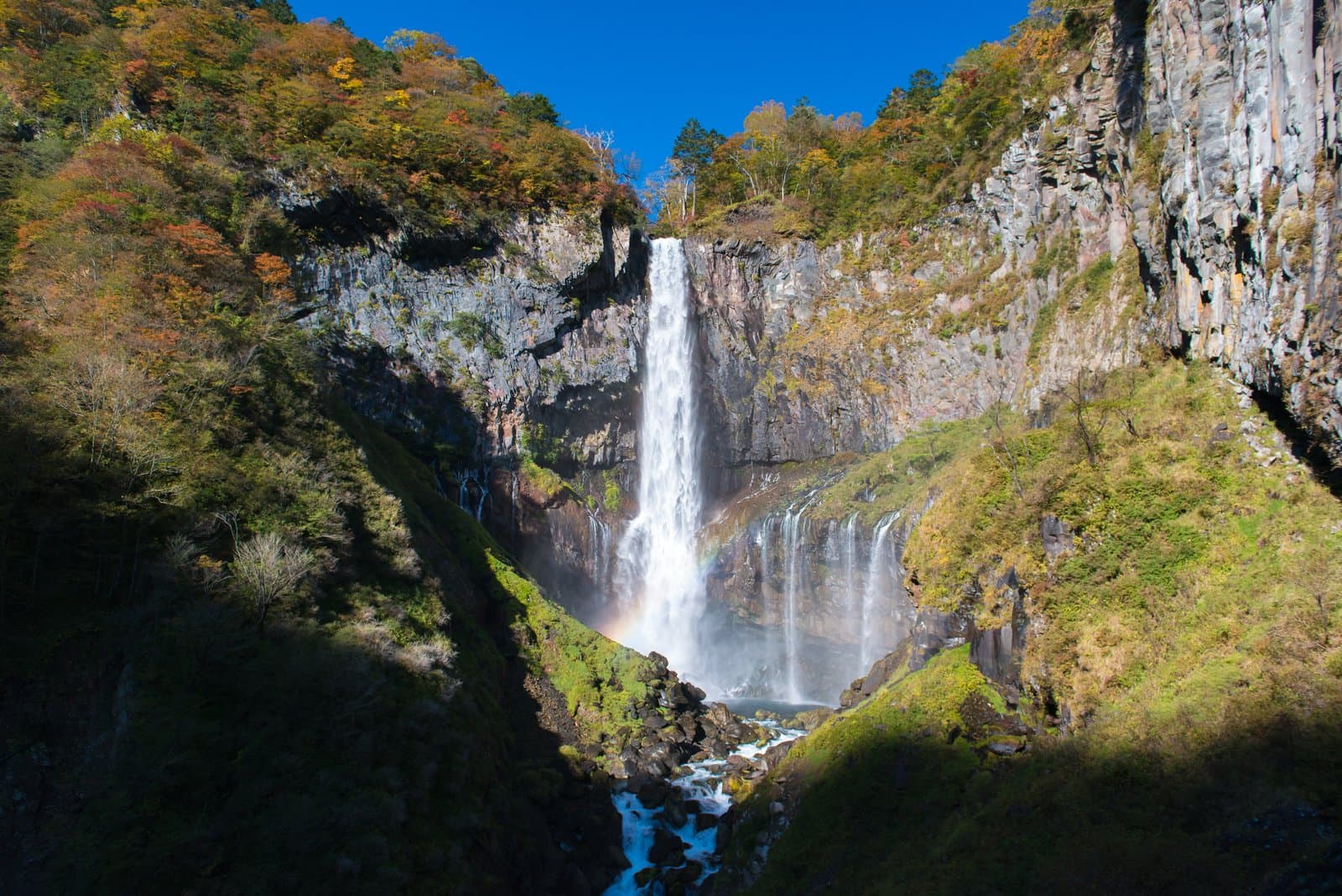A tall waterfall dropping down a sheer cliff between autumn-coloured hills under a blue sky