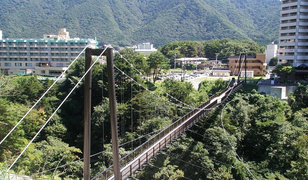A long pedestrian suspension bridge over a wooded gorge, with hot-spring hotels and green mountains beyond
