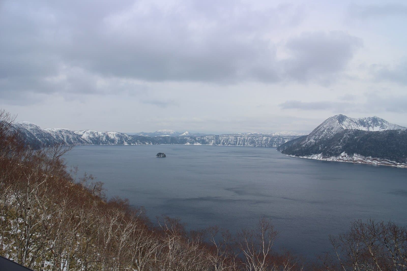 A wide deep-blue lake with a small island, surrounded by snowy cliffs and a snow-capped peak on the right