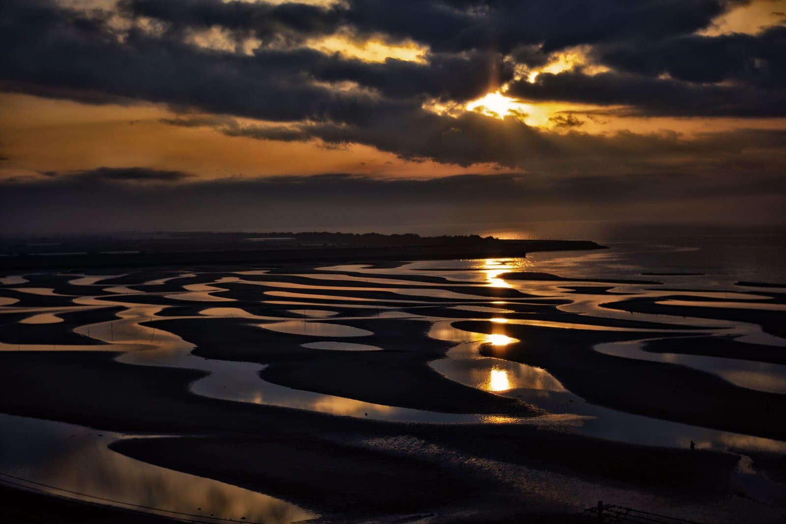Patterns of water on a tidal flat reflecting the setting sun under dark clouds