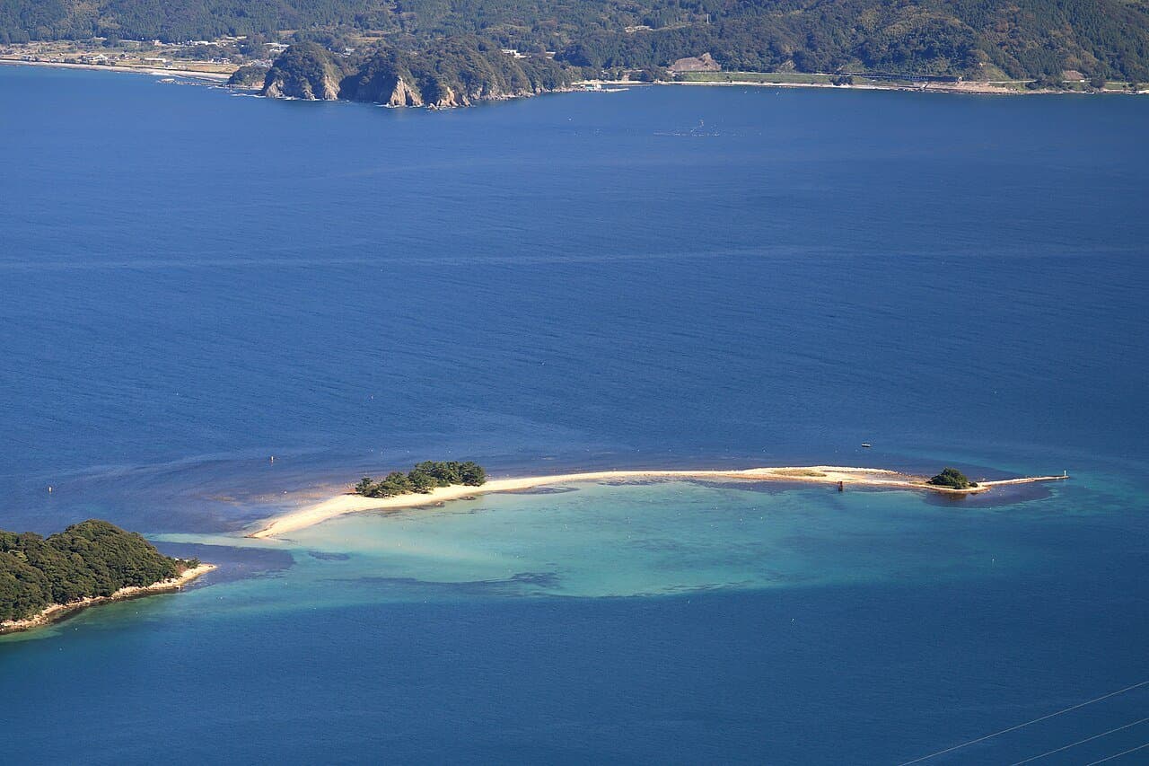 A long white sandbar with two small wooded islets in clear turquoise and deep blue sea, seen from high above