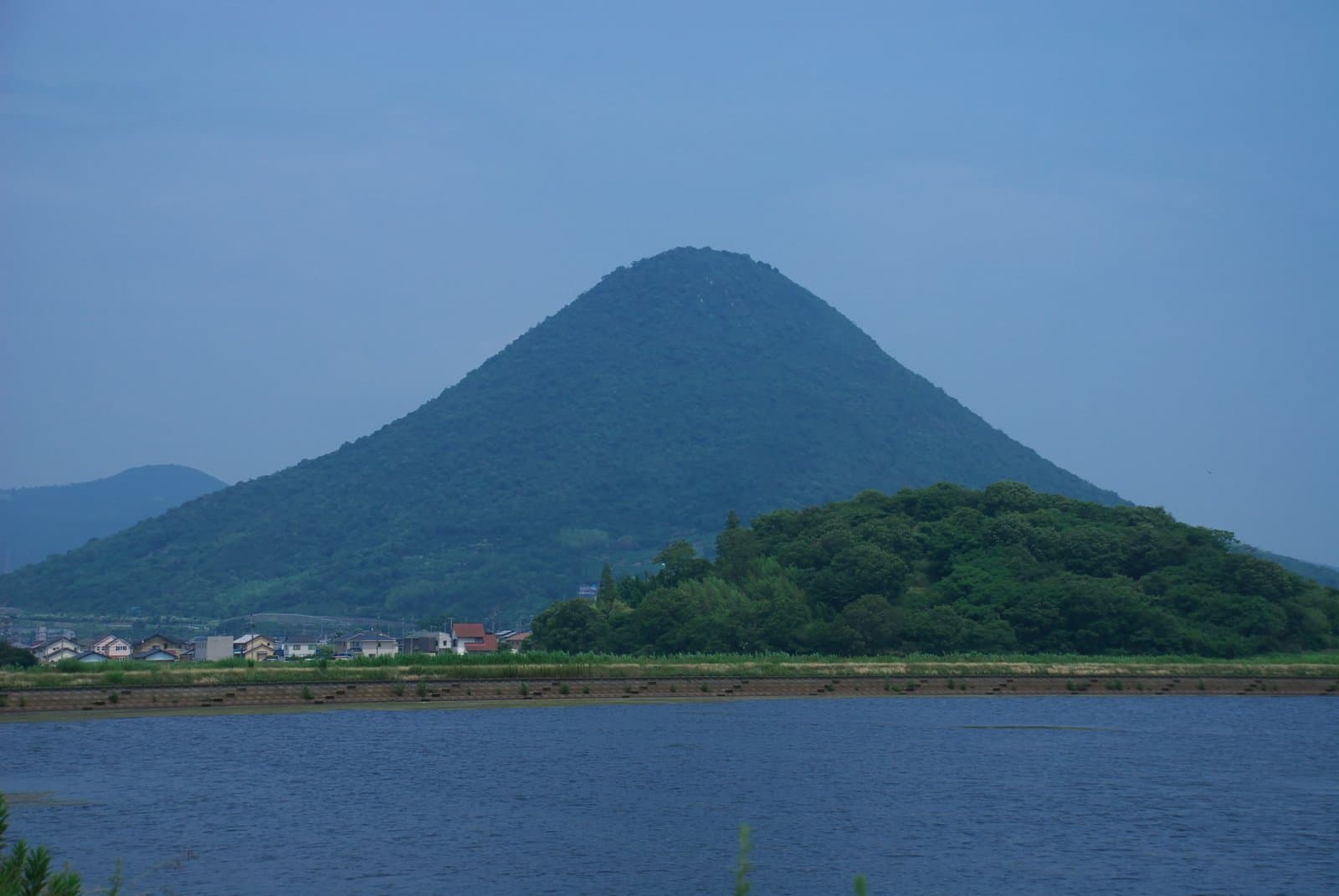 A symmetrical cone-shaped green mountain rising behind a low wooded hill and a stretch of water