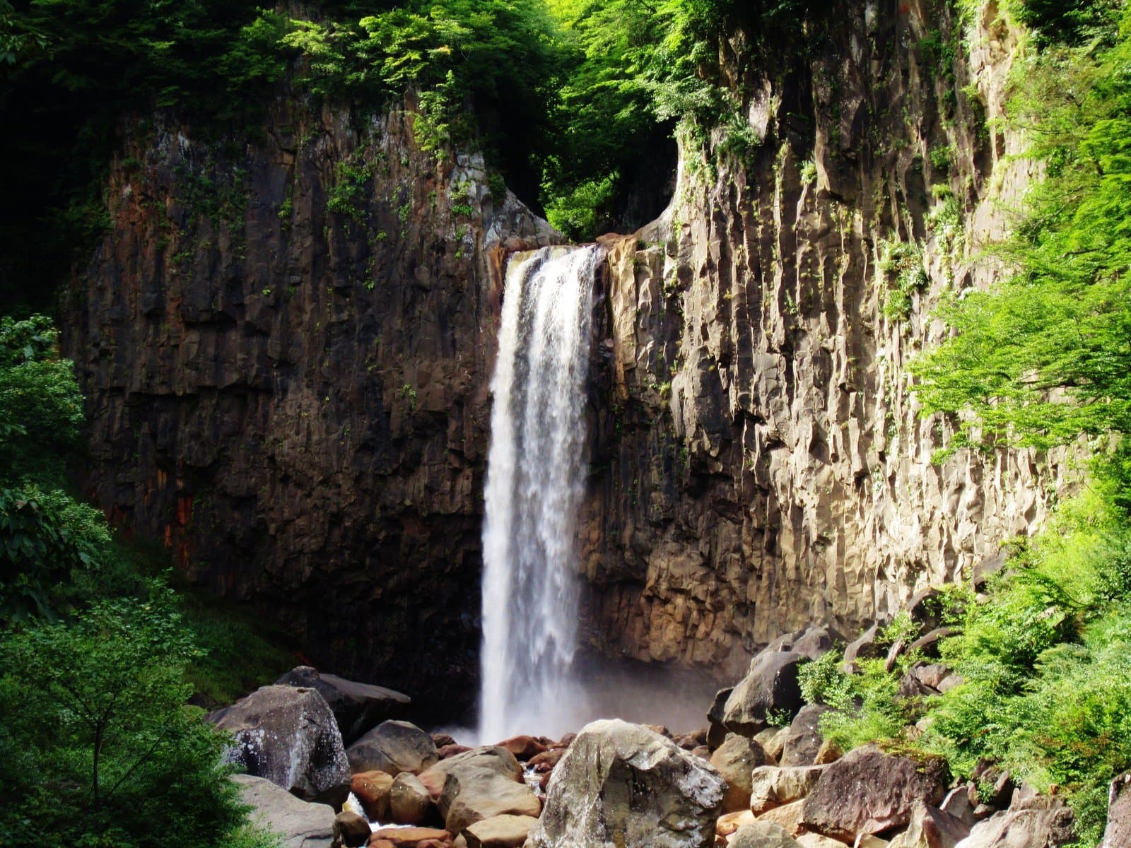 A white waterfall plunging between cliffs of columnar basalt into a boulder-strewn pool, with green foliage