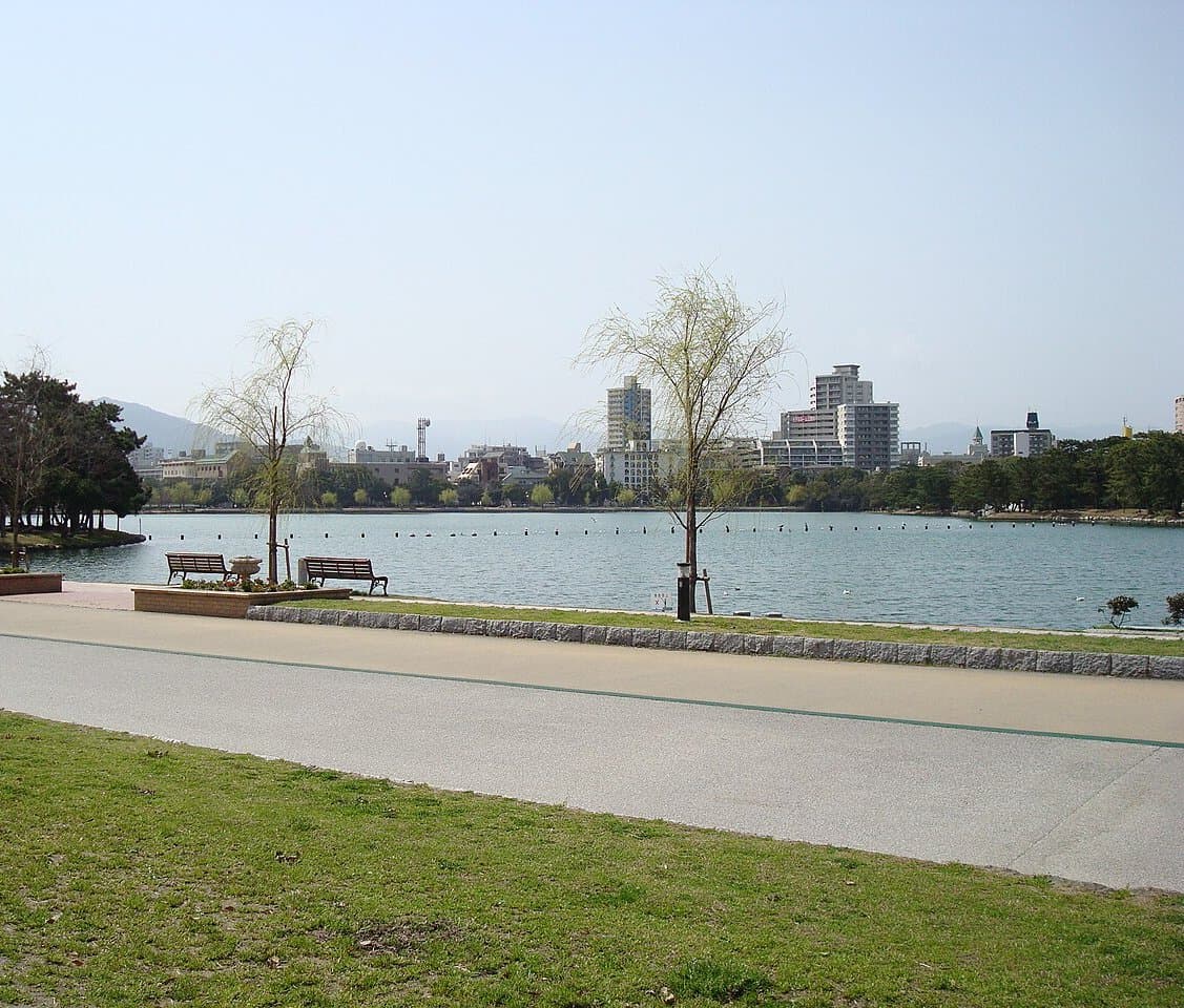 A wide pond with a lakeside path, benches and young willow trees, with city buildings across the water