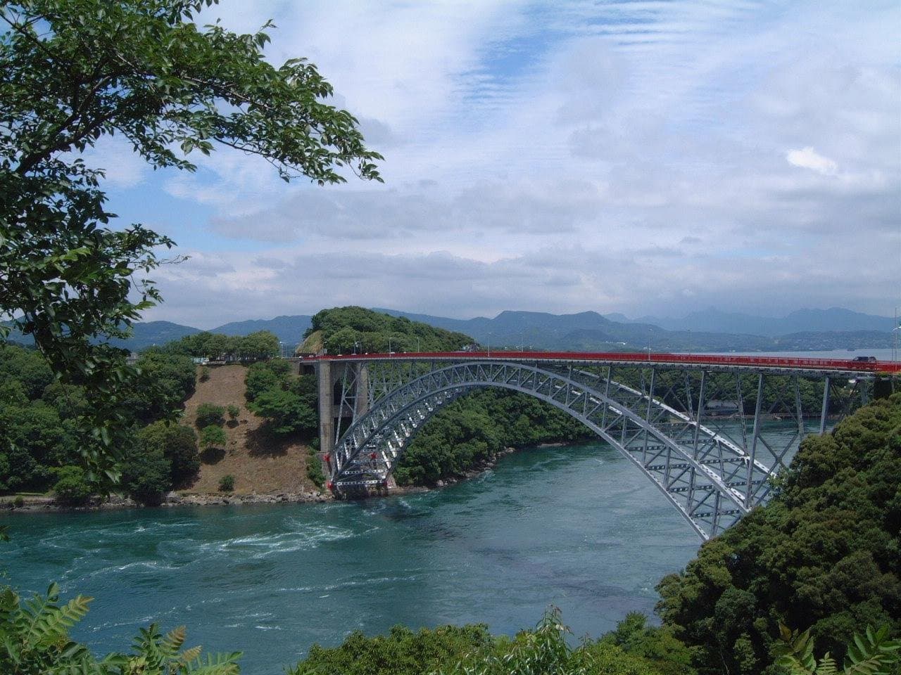 A steel arch bridge over a fast-flowing blue strait between wooded shores, with mountains beyond