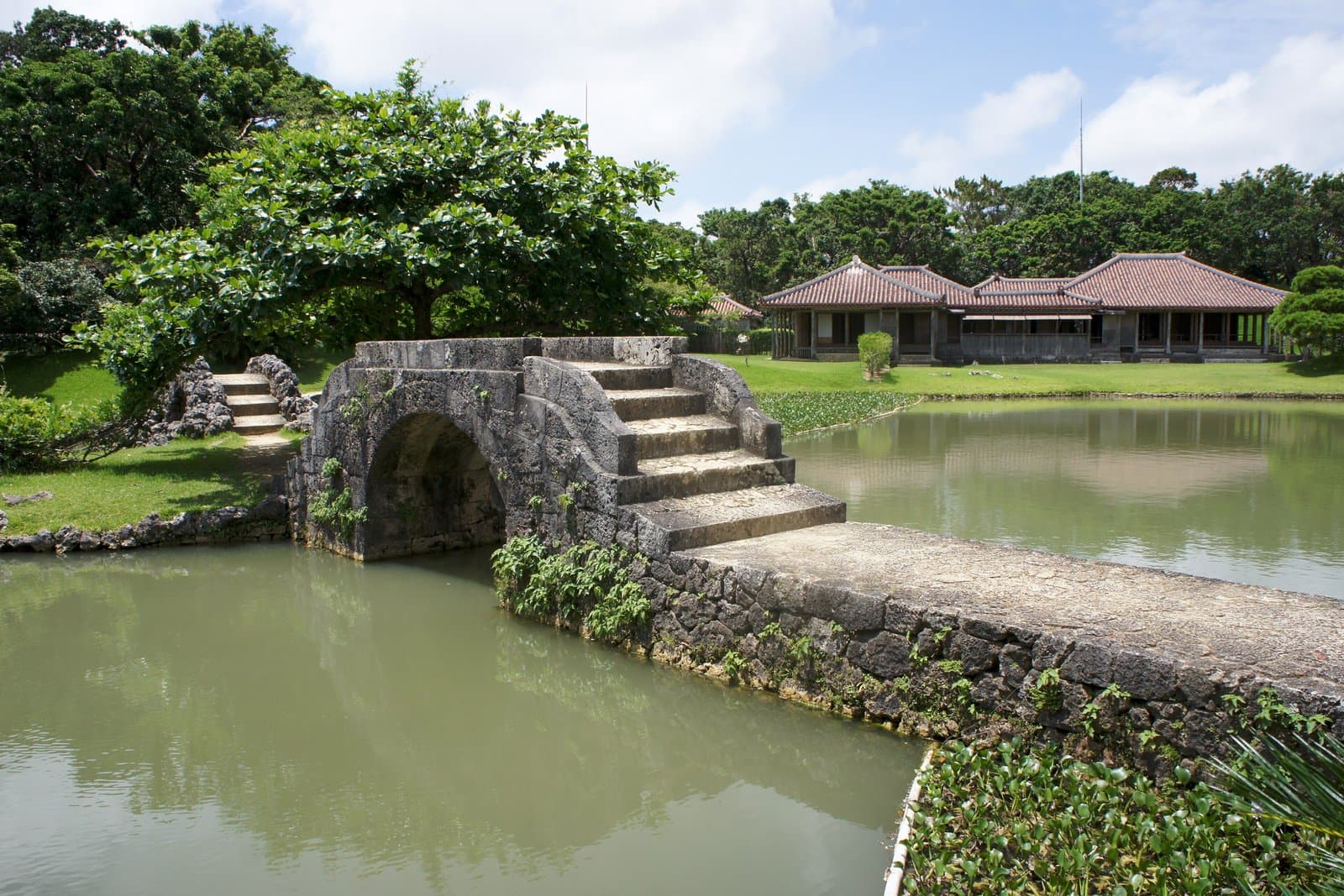 池にかかる石のアーチ橋と、木々の間の赤瓦の屋敷