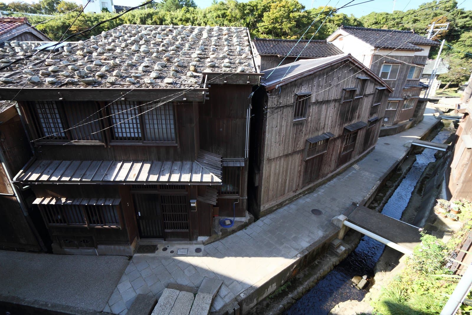 Old wooden houses with stone-weighted roofs along a narrow lane and a small stream