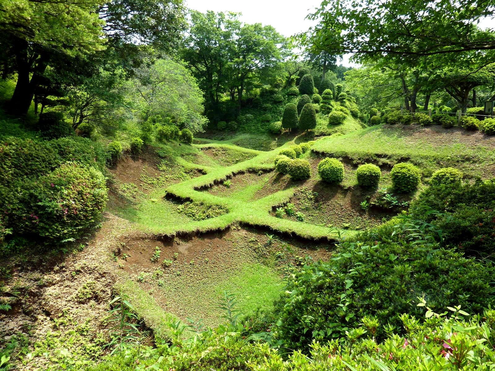 Grassy earthen moats divided into a grid by low ridges, surrounded by trees and clipped shrubs