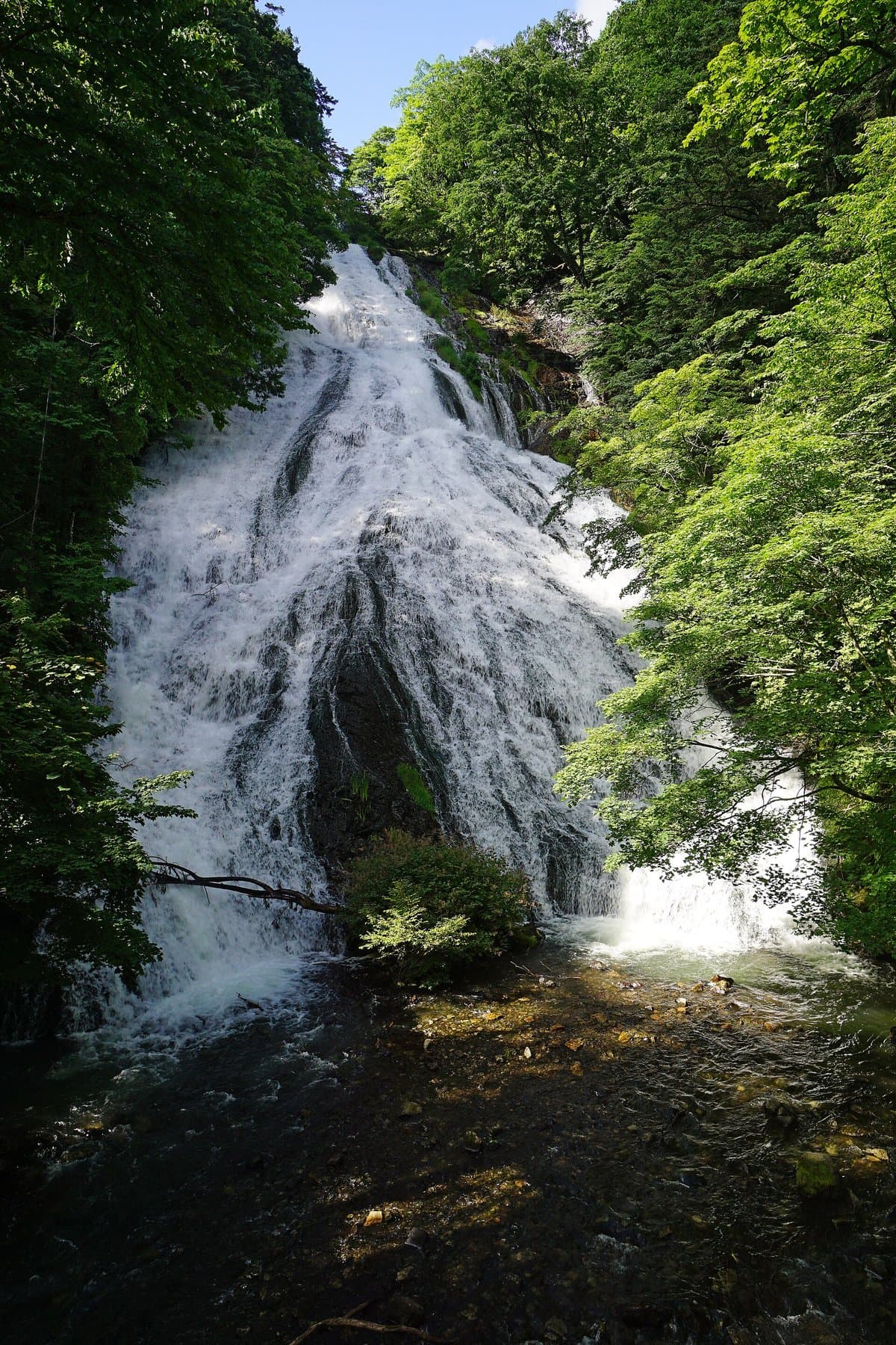 A broad white waterfall sliding down a steep rock face into a pool, framed by fresh green trees